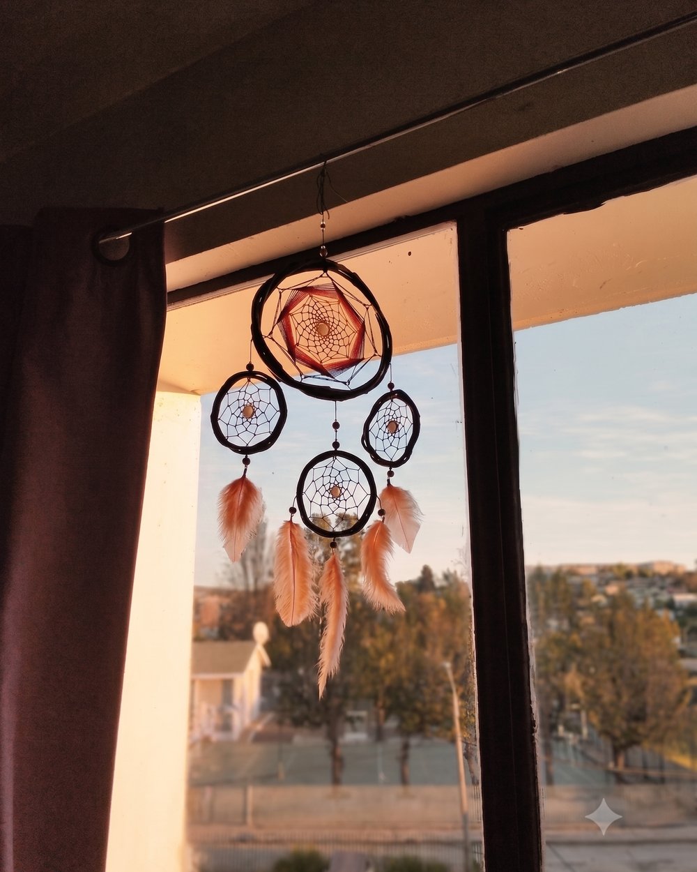 Atrapasueños de madera y plumas colgado frente a una ventana, con cielo despejado al fondo.